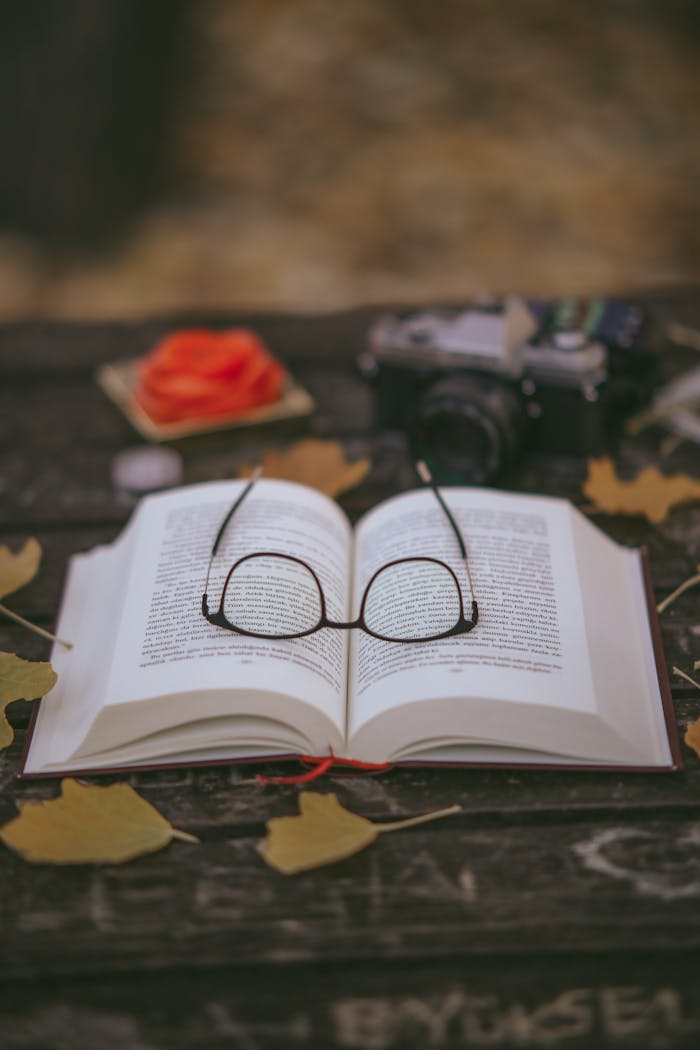An open book with reading glasses on a wooden table surrounded by autumn leaves.