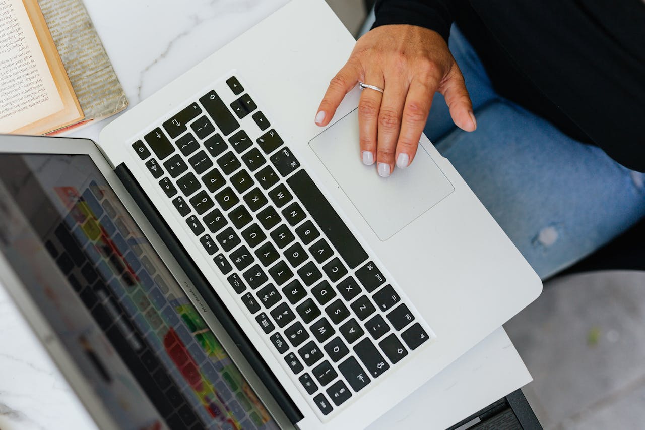 A person working on a laptop at a desk with an open book nearby, viewed from above.